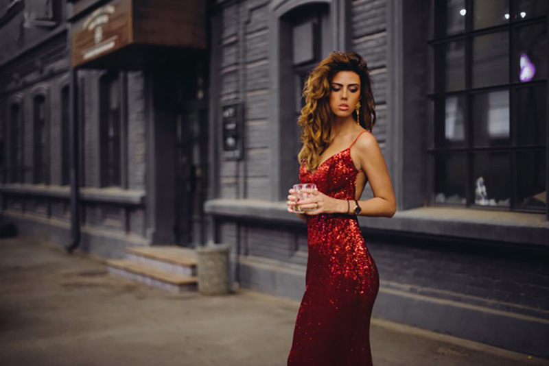 Stylish lady in a red glitter dress holding a glass in front of a grey building